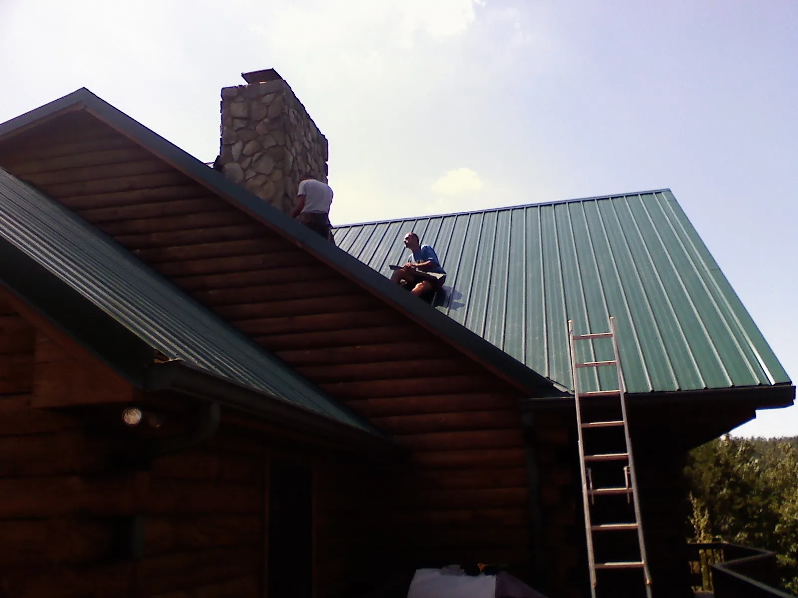 Roofers installing green metal panels beside a stone chimney on a log home in Ventnor City